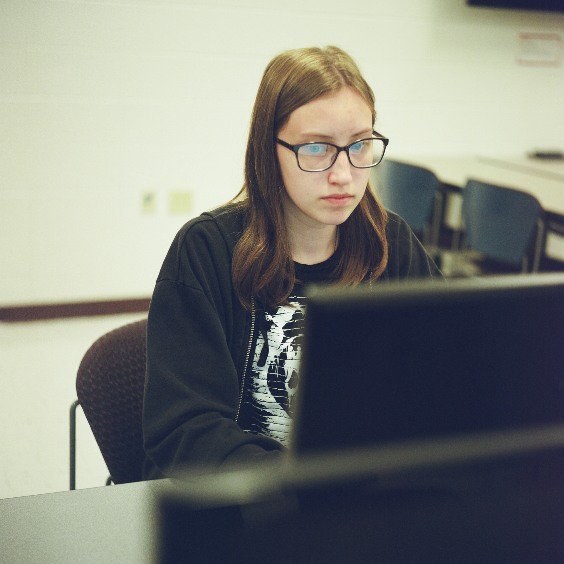 Dellah Hall sits and faces an out-of-focus monitor in the foreground, in a room with out-of-focus plastic stackable chairs facing a table in the background.