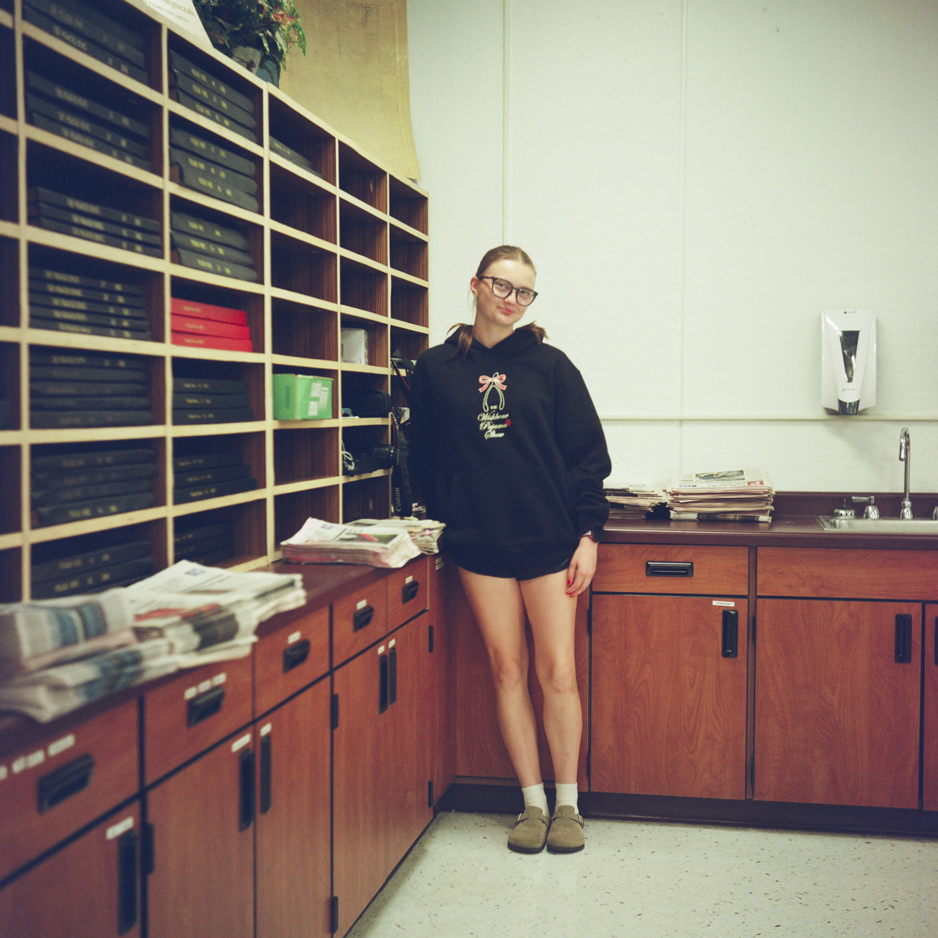 Amelia Lytie poses for a portrait while standing next to a row of wood cabinets and a row of storage shelves filled with bound volumes of newspapers in a room with a vinyl panel floor and painted concrete block wall.