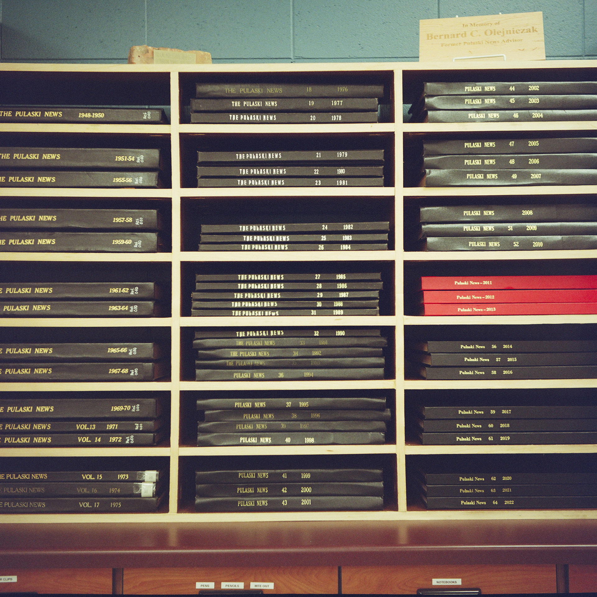 Multiple bound volumes of a newspaper are stacked on their sides in more than 20 slots of a shelf on top of a wood desk and in front of a painted concrete block wall.