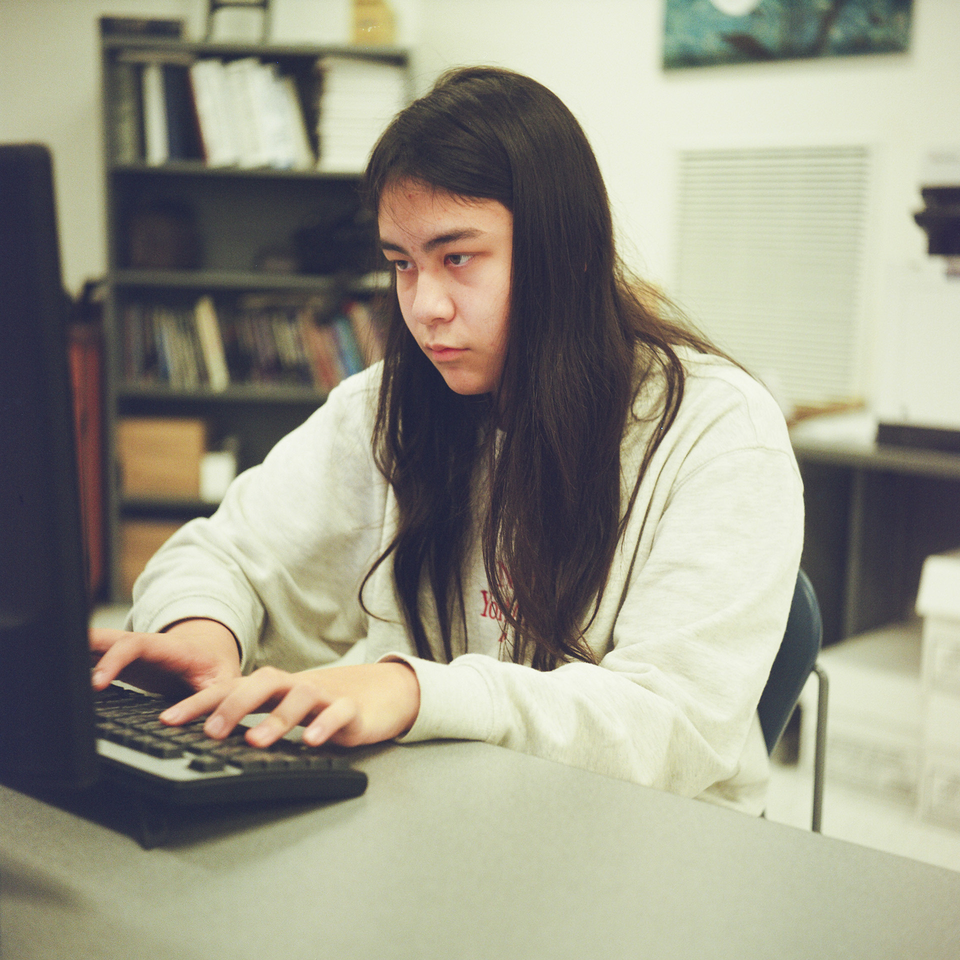 Neville Nguyen types on a keyboard while facing a computer monitor while seated in a room with an out-of-focus bookshelf filled with books and a table with boxes underneath it in the background.