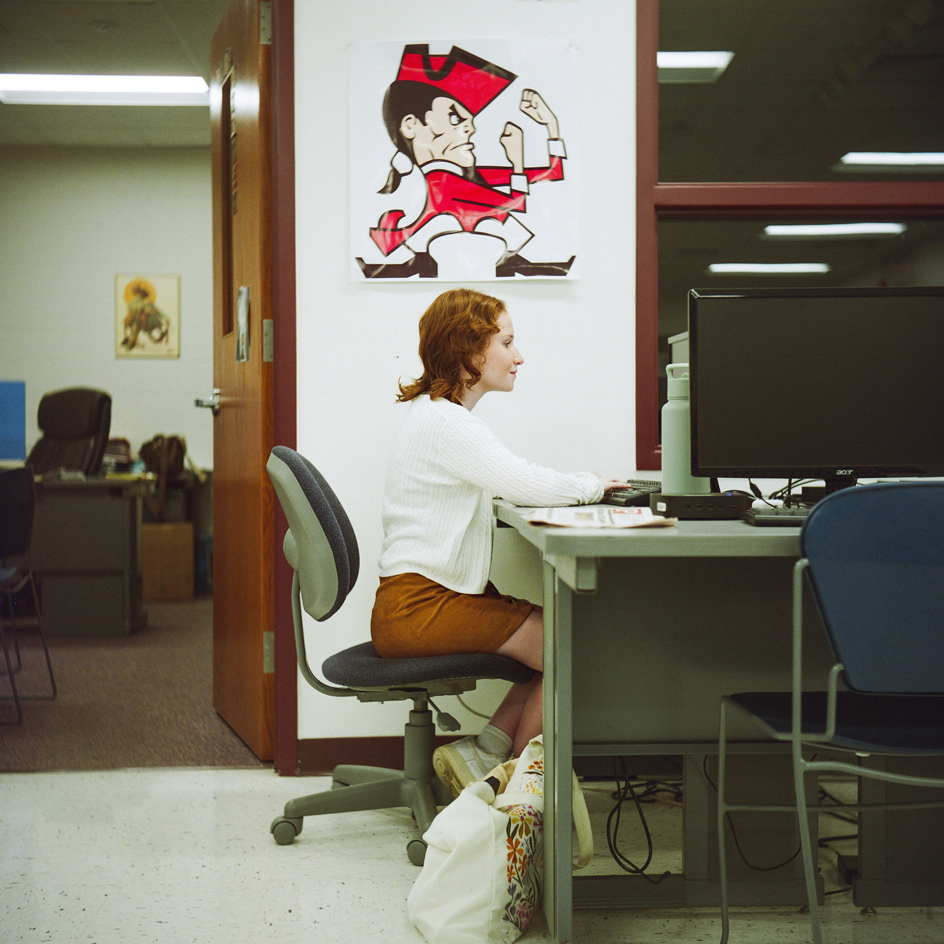 Madelyn Rybak sits in a rolling office chair at a table with multiple computer monitors, papers and a water bottle on its surface, in a room with a decal of a "Red Raiders" mascot on a wall between an open wood door leading to another room and a window with multiple panels.