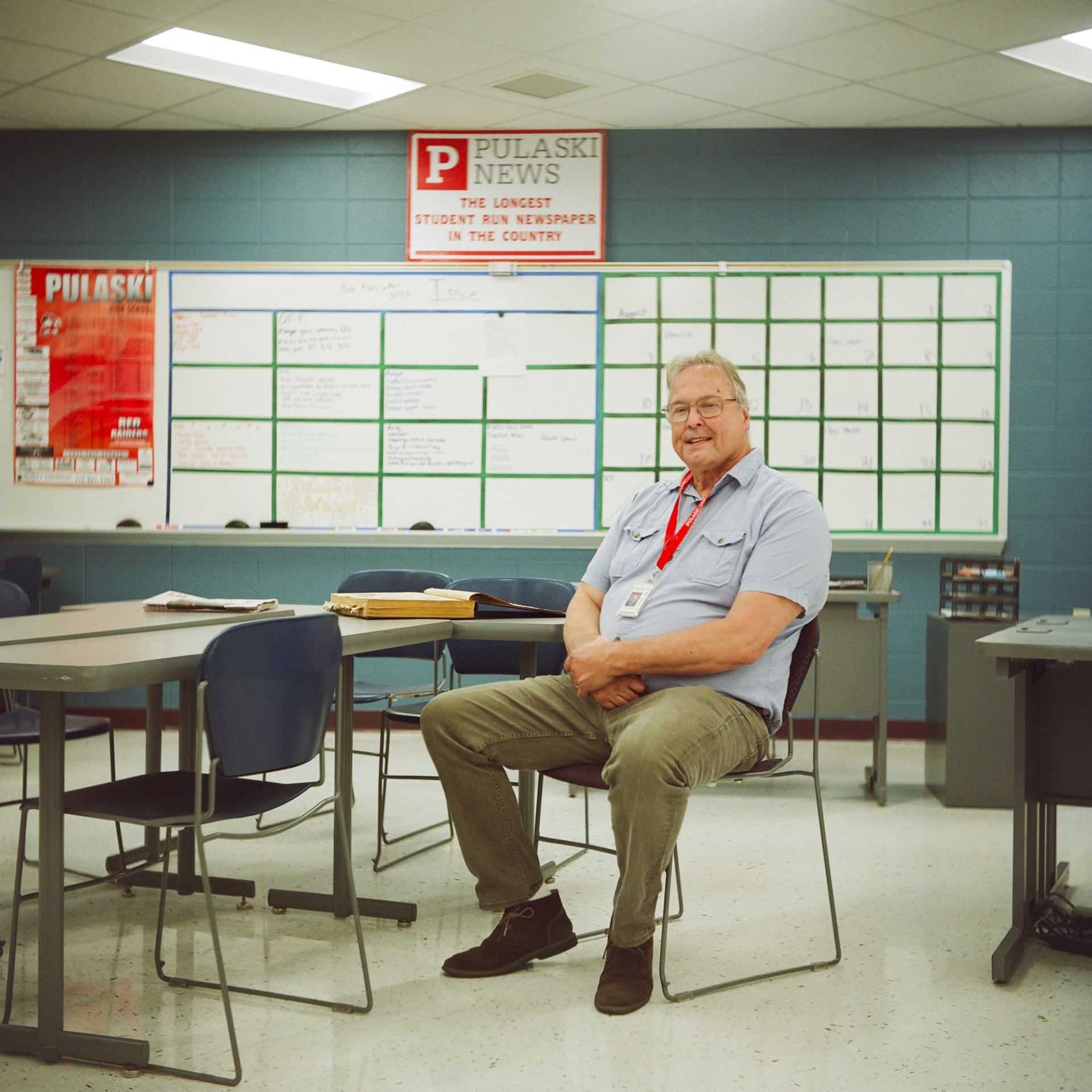 Bob Van Enkenvoort poses for a portrait while sitting in a plastic stackable chair in a room with other chairs arranged around tables, with multiple whiteboards divided with painters tape into smaller segments and a sign reading "Pulaski News" and "The Longest Student Run Newspaper in the Country" mounted on a painted concrete block wall, in a room with a vinyl panel floor and fluorescent lights in a drop panel ceiling.