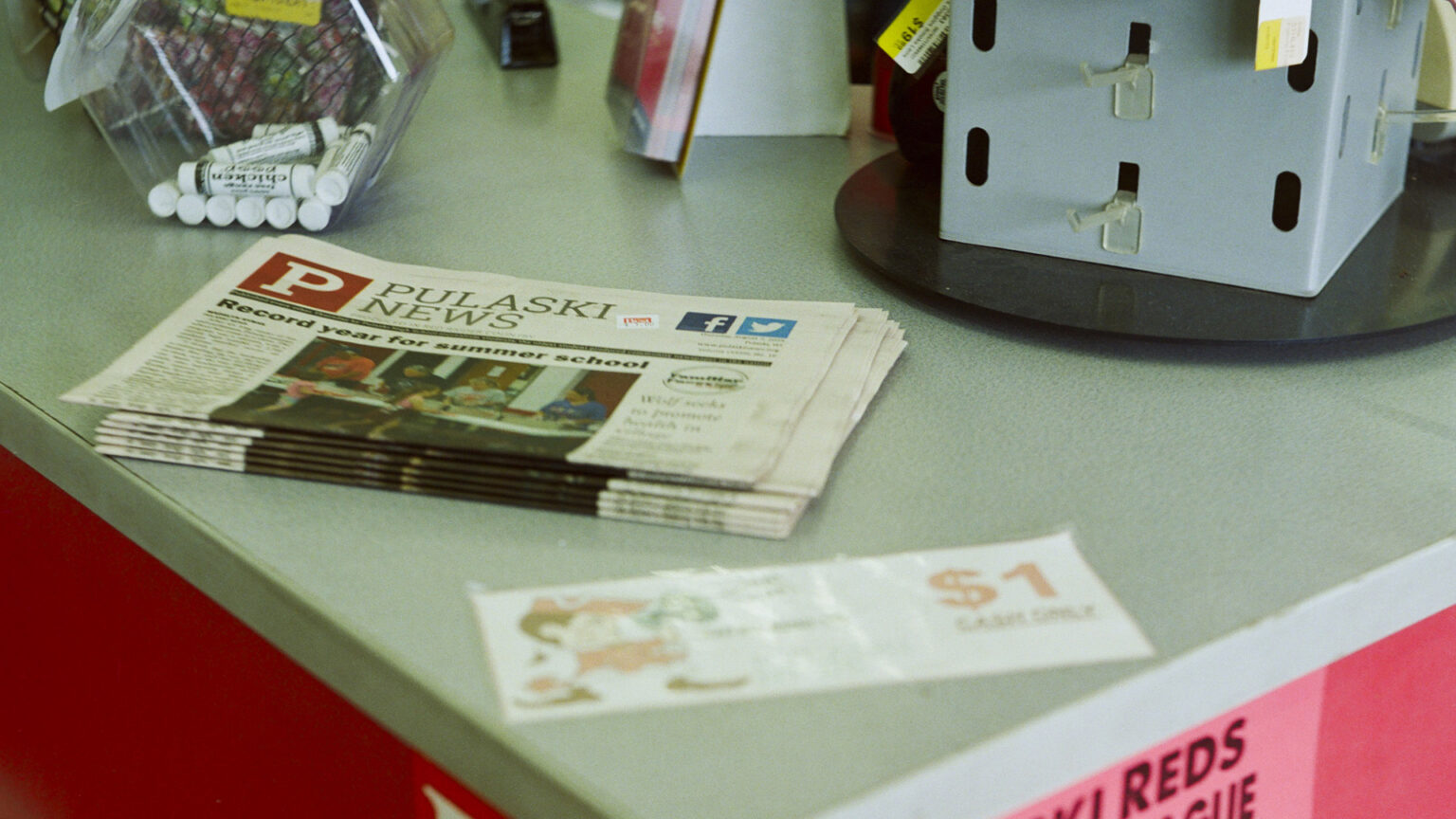 A stack of six newspapers titled Pulaski News with a P logo int he upper left corner sit on a counter top next to a candy jar containing lip balm and a revolving stand with empty hooks. A stack of six newspapers titled Pulaski News with a P logo int he upper left corner sit on a counter top next to a candy jar containing lip balm and a revolving stand with empty hooks.