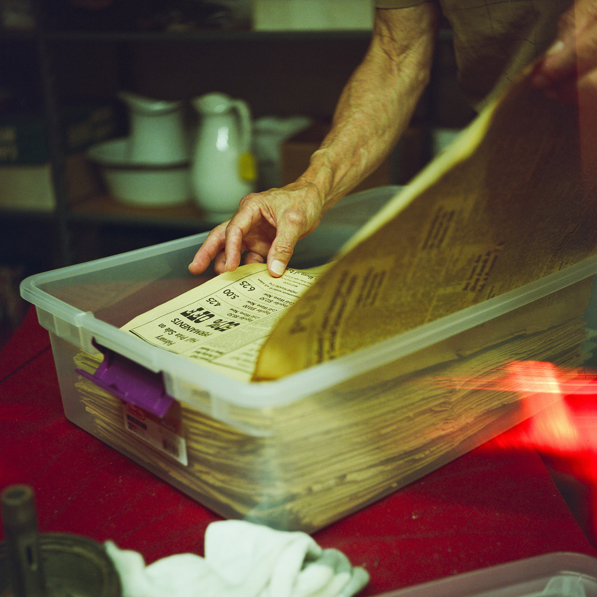 Steve Peplinski's left hand holds the front page of a newspaper while his right hand lifts its next page from the top of a stack of aging newspapers inside a plastic storage bin sitting on the surface of a table, with out-of-focus pitchers and basins on a shelf in the background.