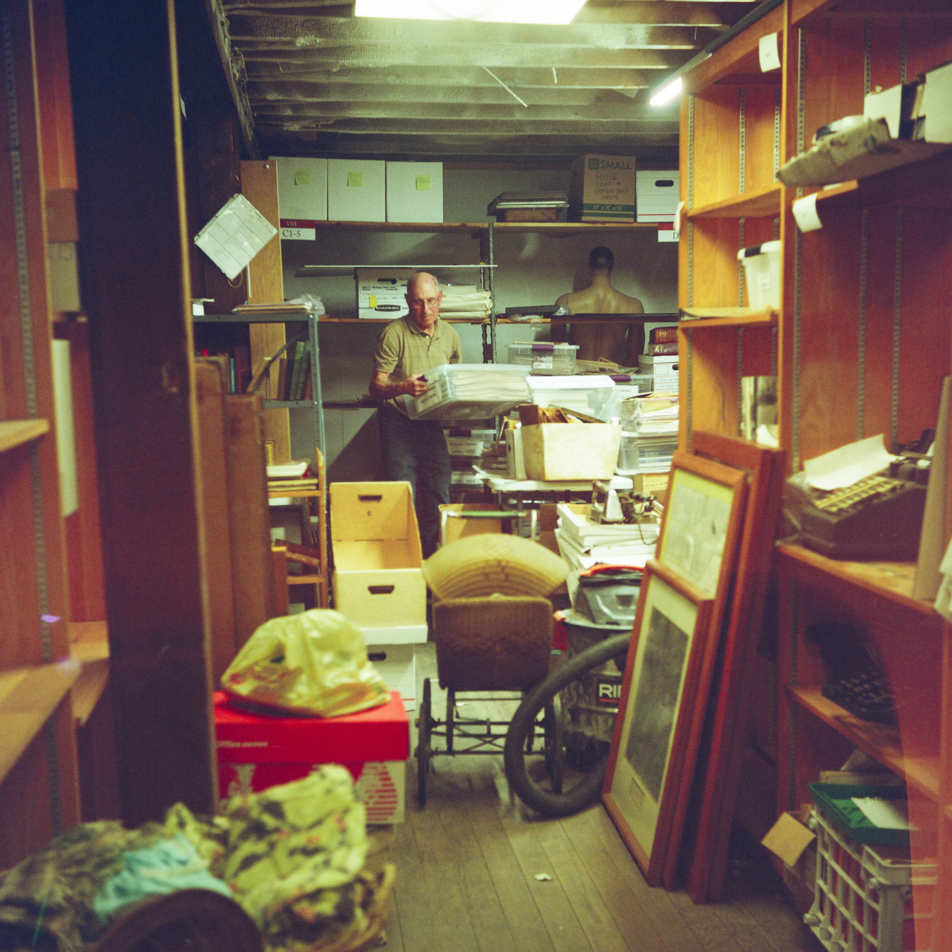 Steve Peplinski carries a plastic box filled with newspapers while walking among stacks of items filling a storage space with wood and metal shelves in a room with a wood floor and wood-beam ceiling.