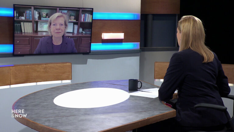 Frederica Freyberg sits at a desk on the Here & Now set and faces a video monitor showing an image of Tammy Baldwin.