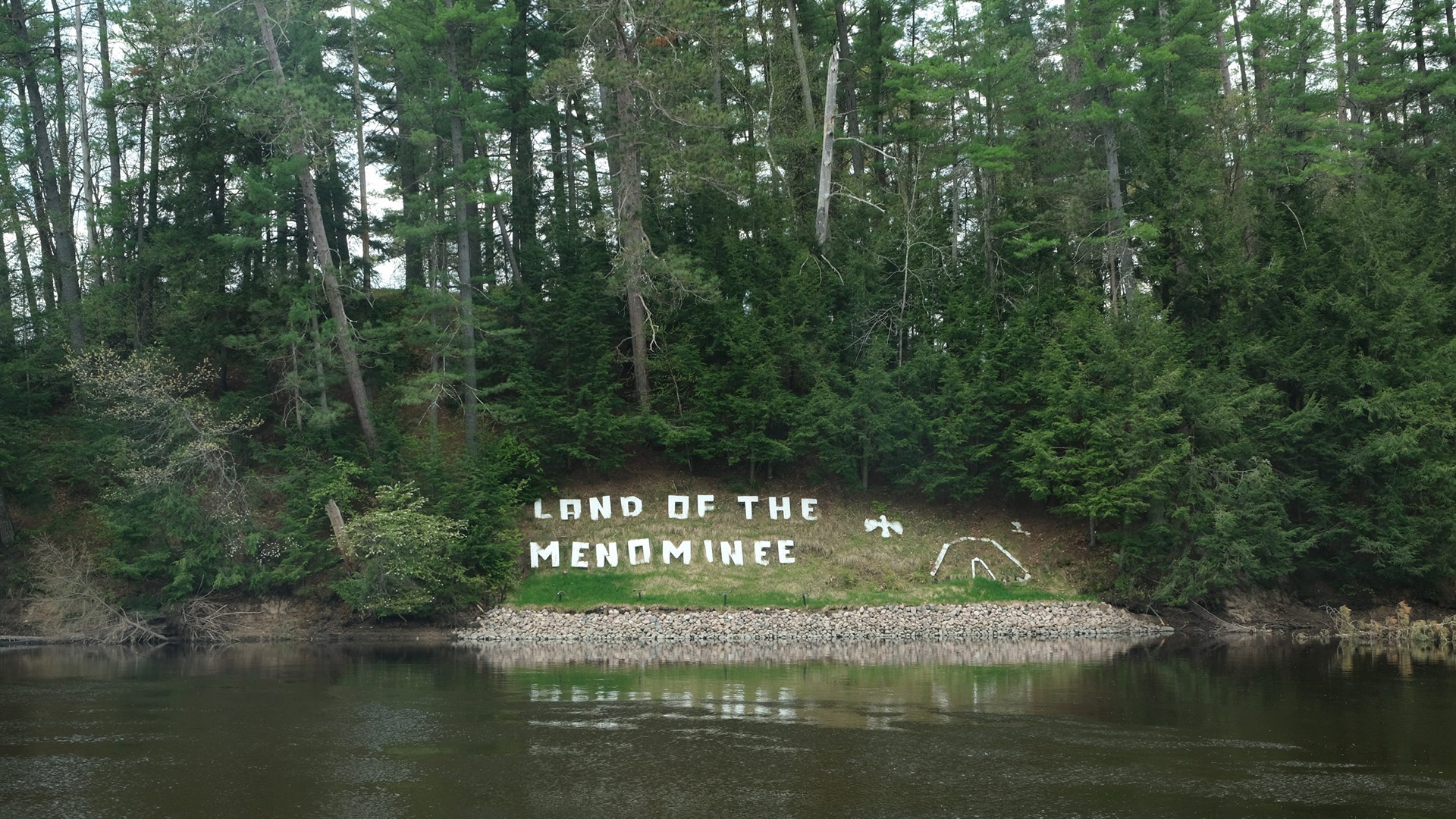 A letter sign reading "Land of the Menominee" with multiple symbols rests at the bottom of a hillside clearing surrounded by trees and above a stone berm along the shore of a body of water.