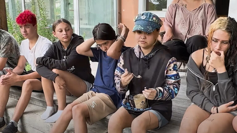 A group of Stockbridge-Munsee children sit in a row on the edge of a wood deck, with one adjusting their hair and another eating a container of gelato while holding a microphone in their left hand, with an adult seated on a chair behind them in front of the windows of a building.