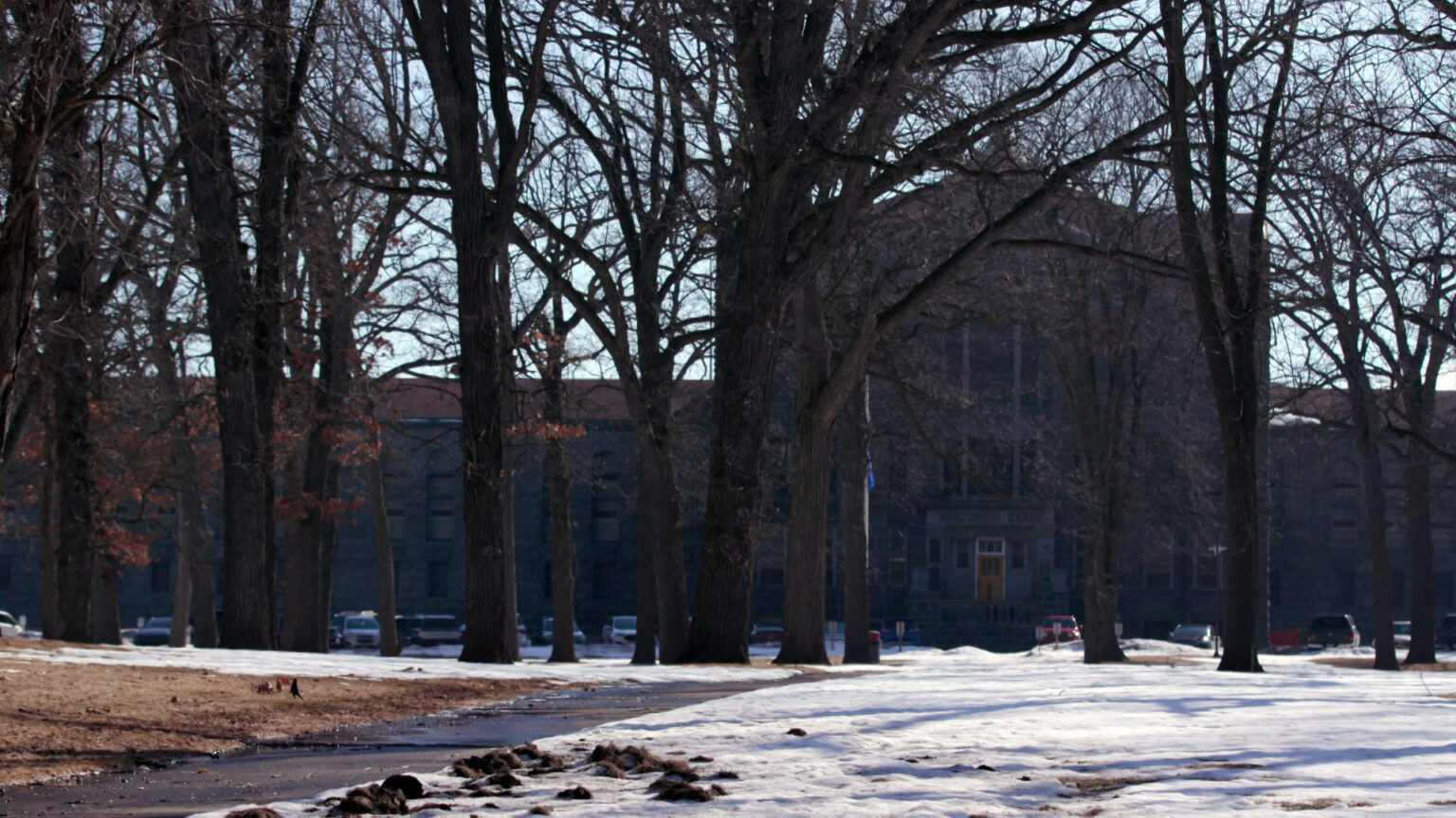 Mature leafless trees stand above a snow-covered lawn, with a concrete sidewalk leading toward a parking lot with multiple vehicles parked in front of a multi-story masonry building with a central structure topped by a pillar-supported pediment. Mature leafless trees stand above a snow-covered lawn, with a concrete sidewalk leading toward a parking lot with multiple vehicles parked in front of a multi-story masonry building with a central structure topped by a pillar-supported pediment.