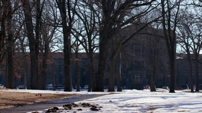 Mature leafless trees stand above a snow-covered lawn, with a concrete sidewalk leading toward a parking lot with multiple vehicles parked in front of a multi-story masonry building with a central structure topped by a pillar-supported pediment. Mature leafless trees stand above a snow-covered lawn, with a concrete sidewalk leading toward a parking lot with multiple vehicles parked in front of a multi-story masonry building with a central structure topped by a pillar-supported pediment.