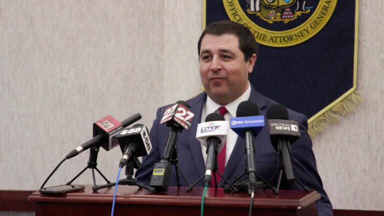 Josh Kaul speaks while standing behind a wood podium with six microphones with the flags of different media organizations standing on tripods in its surface, in a room with a cloth banner showing the seal of Wisconsin's Office of the Attorney General mounted on a wall in the background.