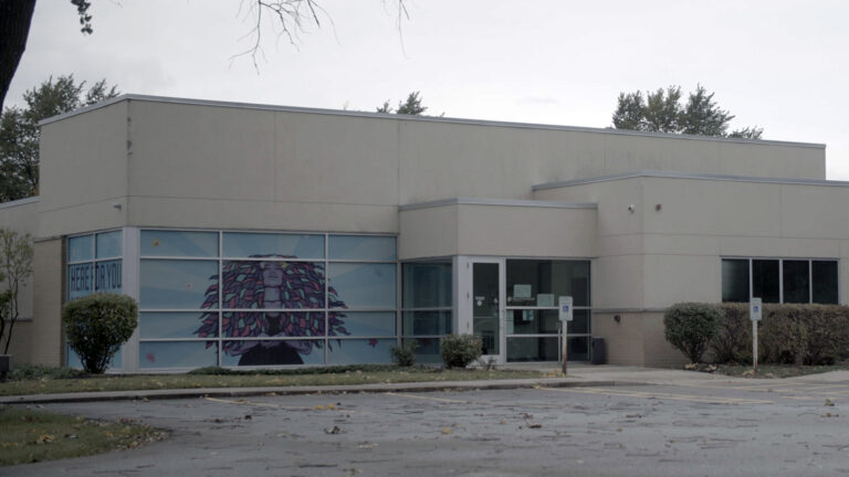 An empty parking lot with leaves and branches on its asphalt surface sits in front of a building with a glass-walled entrance and large window at one corner covered by a decal with an illustration of a woman standing at the center of rays of light, with multiple trees behind it in the background.