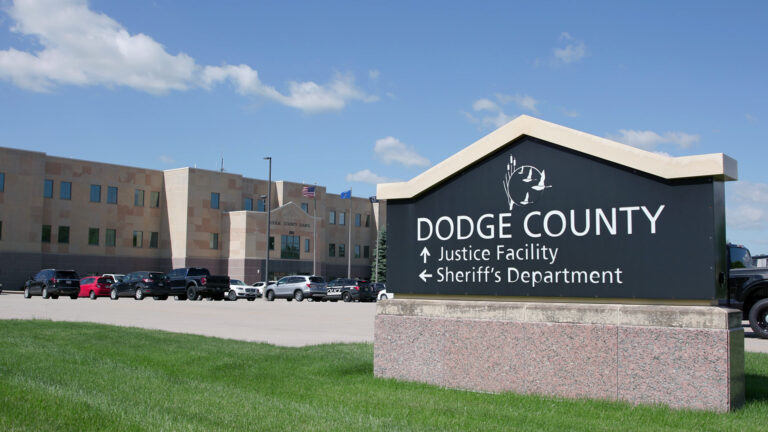A sign in the shape of a peaked roof shows a logo with a flock of flying waterfowl and the words Dodge County, Justice Facility and Sheriff's Department on top of a stone base in a lawn, situated in front of a parking lot filled with vehicles and with a two story stone masonry building with a row of flagpoles flying the U.S., Wisconsin and POW/MIA flags next to its entrance under a partly cloudy sky.