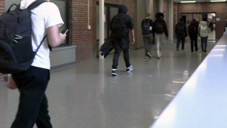 A student with a backpack holds a cellphone in their right hand while walking behind other students in a hallway with a polished floor and brick walls with multiple windows. A student with a backpack holds a cellphone in their right hand while walking behind other students in a hallway with a polished floor and brick walls with multiple windows.