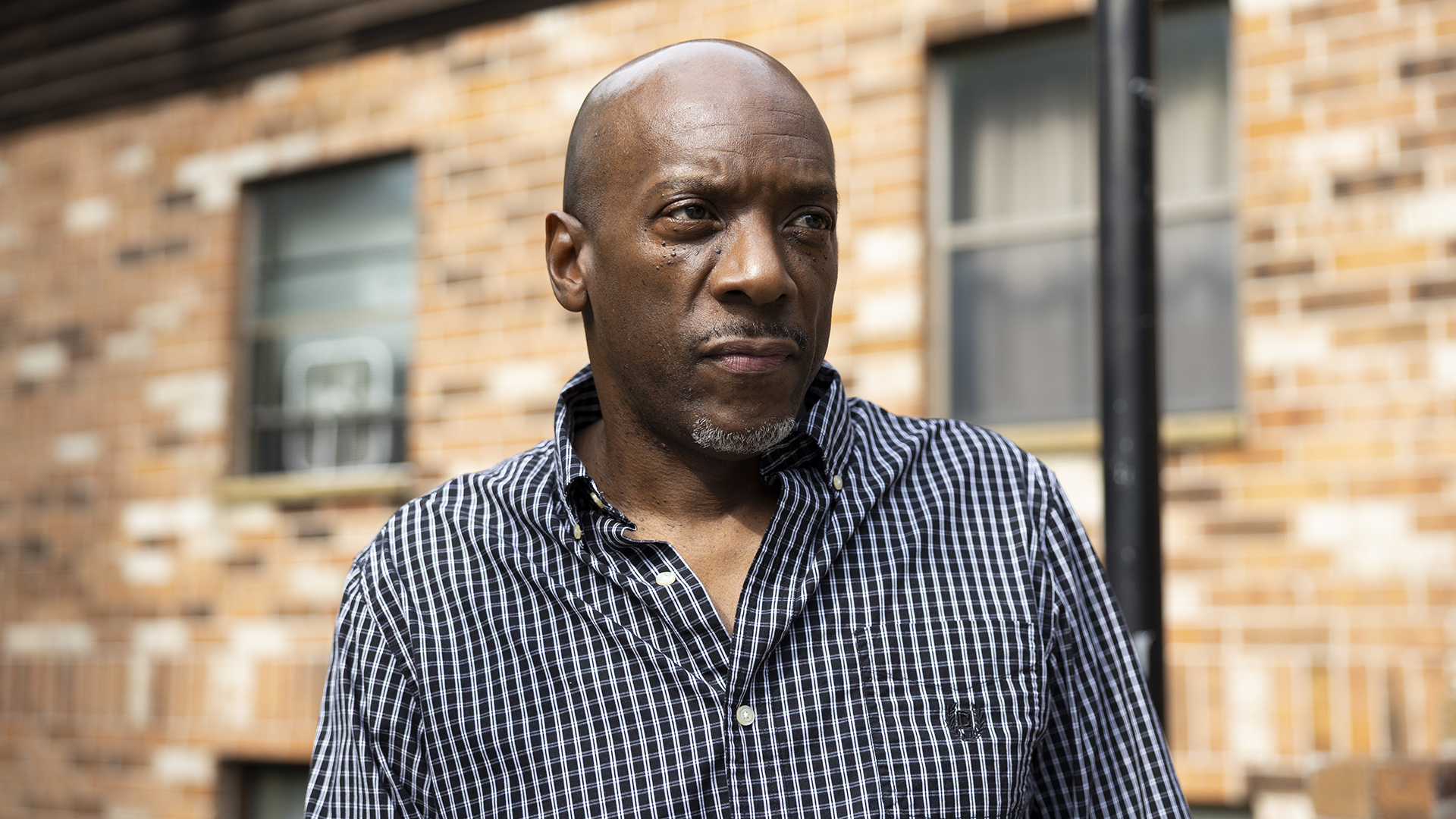 Darnell Price poses for a portrait in half profile while standing in front of a building with a brick wall and multiple windows.