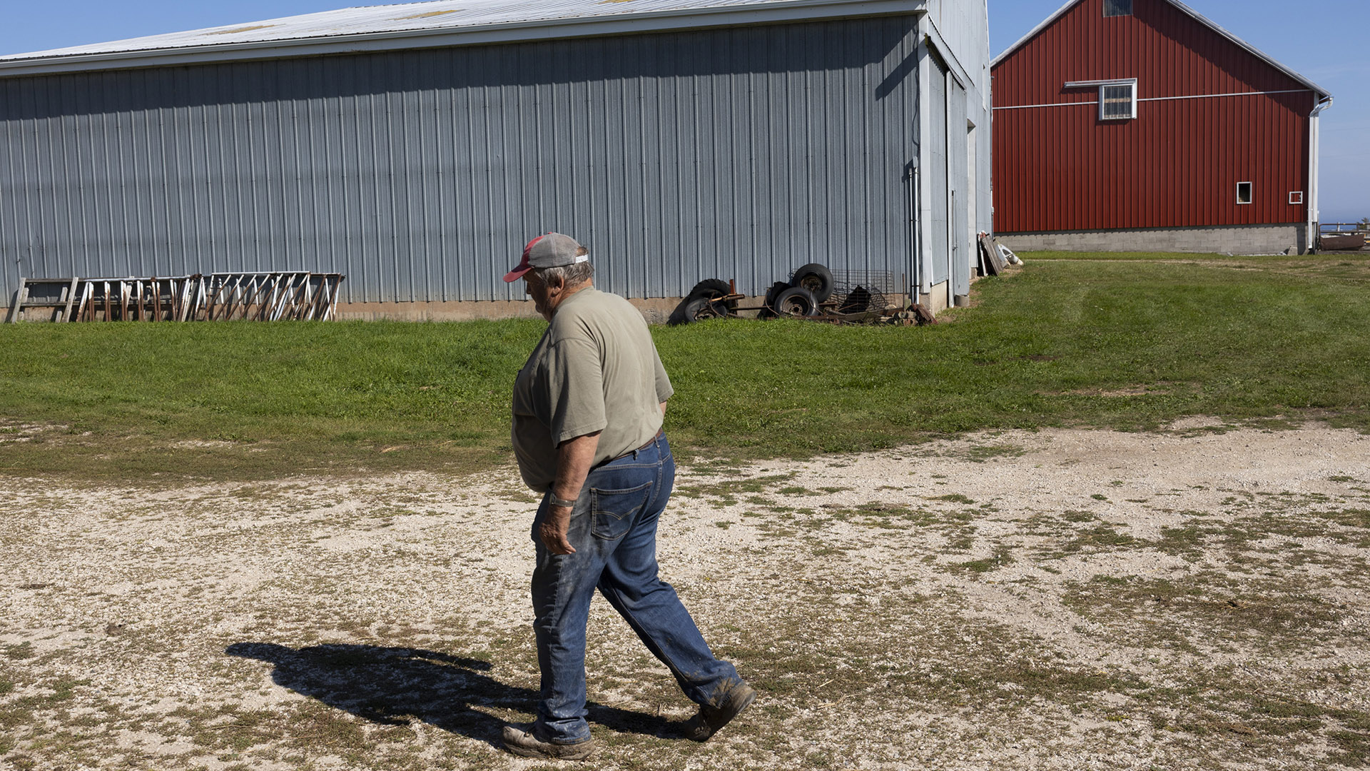 David Hardtke walks on flat graveled area in front of a lawn next to a metal farm building, with another farm building in the background.