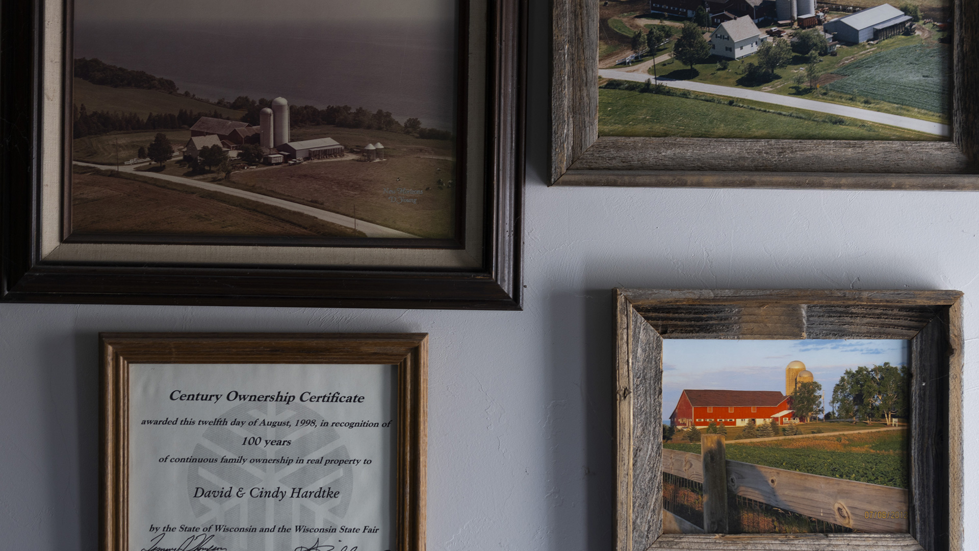 Three framed photos of a farm, two from an aerial perspective and one from a ground-level perspective, as well as a framed certificate with a title reading "Century Ownership Certificate" are mounted on a wall.