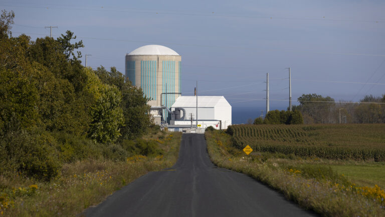 An asphalt road between a wooded area on the left side and farm fields on the right side, with a road sign in the distance reading Stop Ahead, leads toward a cluster of buildings, including a round concrete tower with a low dome, with multiple power towers and lines around the facility and a large body of water in the background.