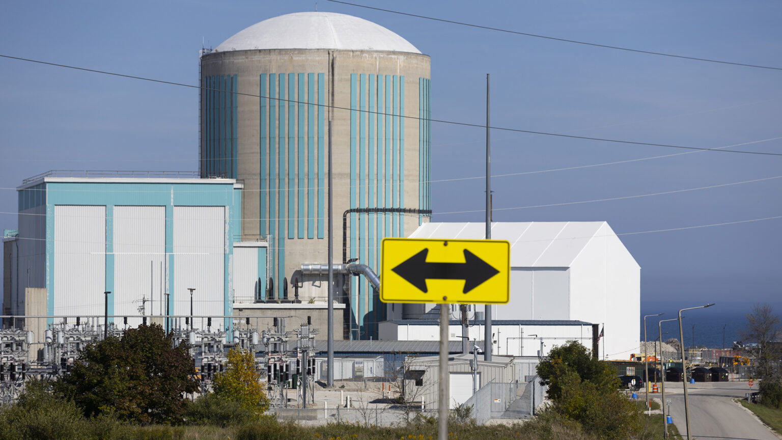 An out-of-focus road sign with arrows pointed in opposite directions stands in the foreground in front of cluster of buildings, including a round concrete tower with a low dome, with multiple power lines around the facility, a road on one side, and a large body of water in the background. An out-of-focus road sign with arrows pointed in opposite directions stands in the foreground in front of cluster of buildings, including a round concrete tower with a low dome, with multiple power lines around the facility, a road on one side, and a large body of water in the background.