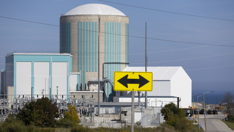An out-of-focus road sign with arrows pointed in opposite directions stands in the foreground in front of cluster of buildings, including a round concrete tower with a low dome, with multiple power lines around the facility, a road on one side, and a large body of water in the background.