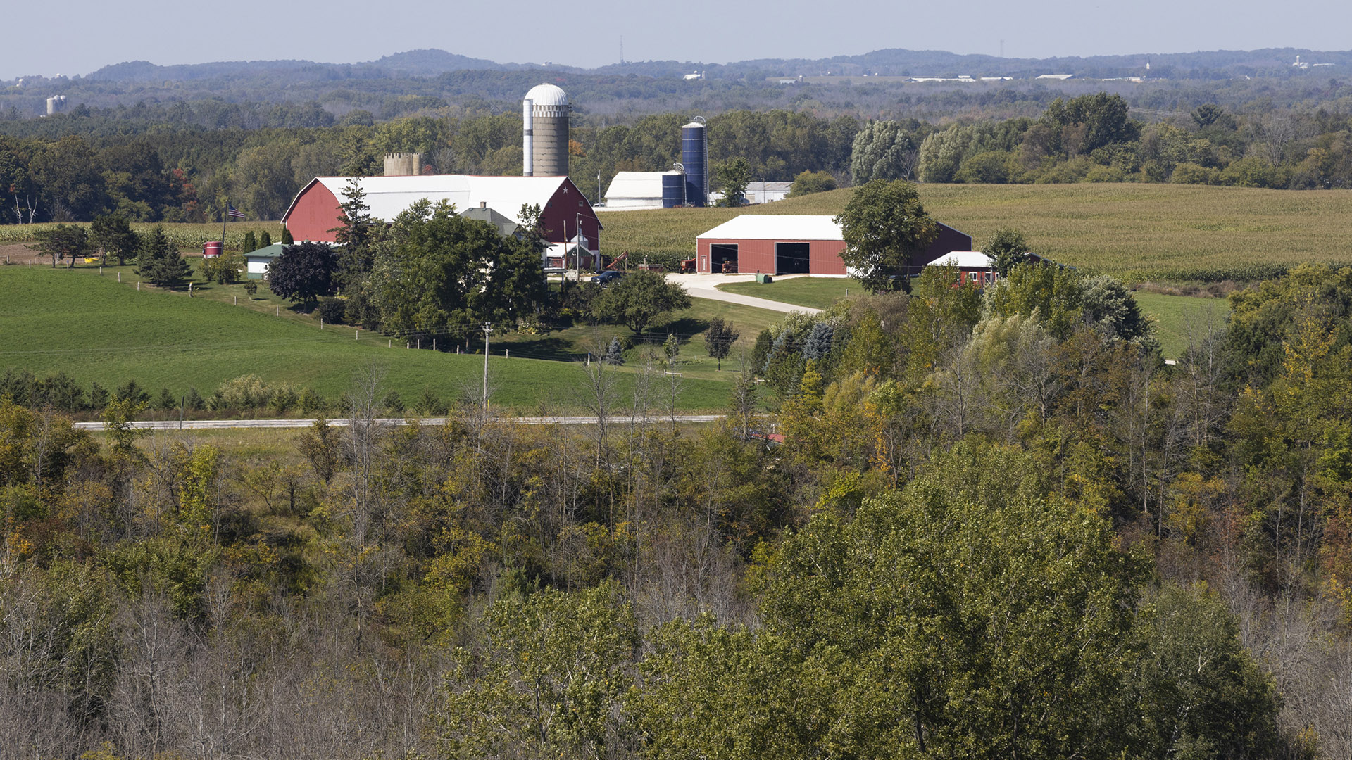 A farm with multiple barns, silos and other buildings stand among trees and fields, with a wooded area in the foreground and more wooded land extending to hills on the horizon.