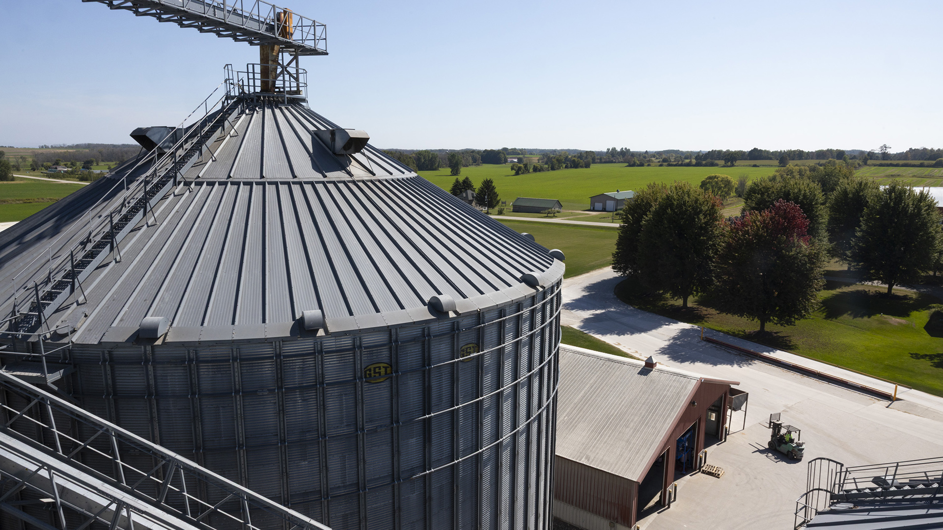 A grain bin with multiple walkways along, above and next to its sloped roof stands over a farm outbuilding with multiple vehicle-sized doors next to a road, with fields and wooded areas in the background.