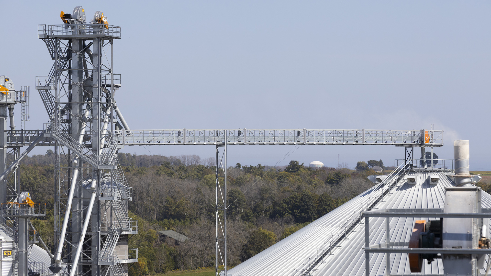 Multiple metal stairways and walkways stand next to the sloped roof of a grain bin, with a wooded area in the background and a round building with a low dome on the horizon.