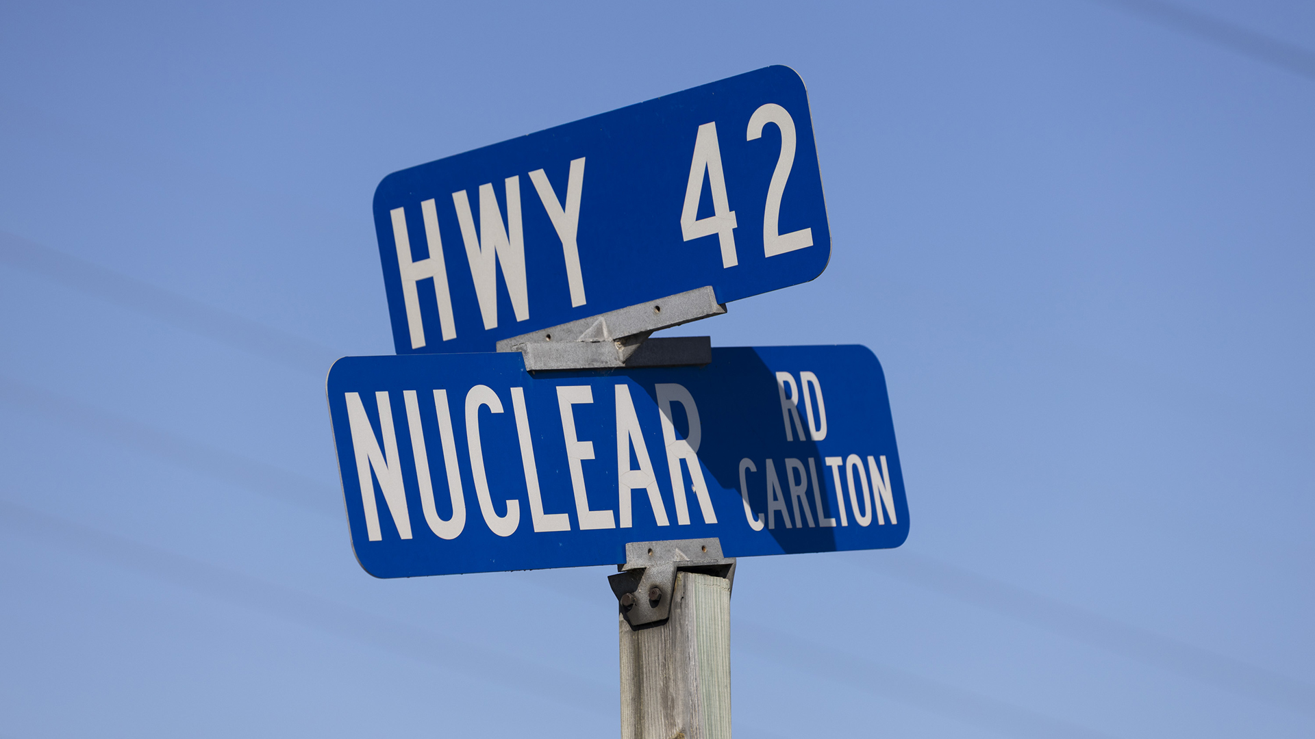 Two metal street signs reading "Hwy 42" and "Nuclear Rd Carlton" are placed in perpendicular directions on top of a square wood stand, with out-of-focus power lines in the background.