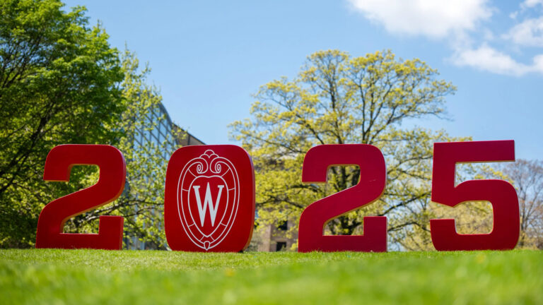 A sign comprised of the numerals 2, 0, 2 and 5, with the UW-Madison W crest logo in the middle of the 0, stands on a lawn, with out-of-focus trees a building in the background.