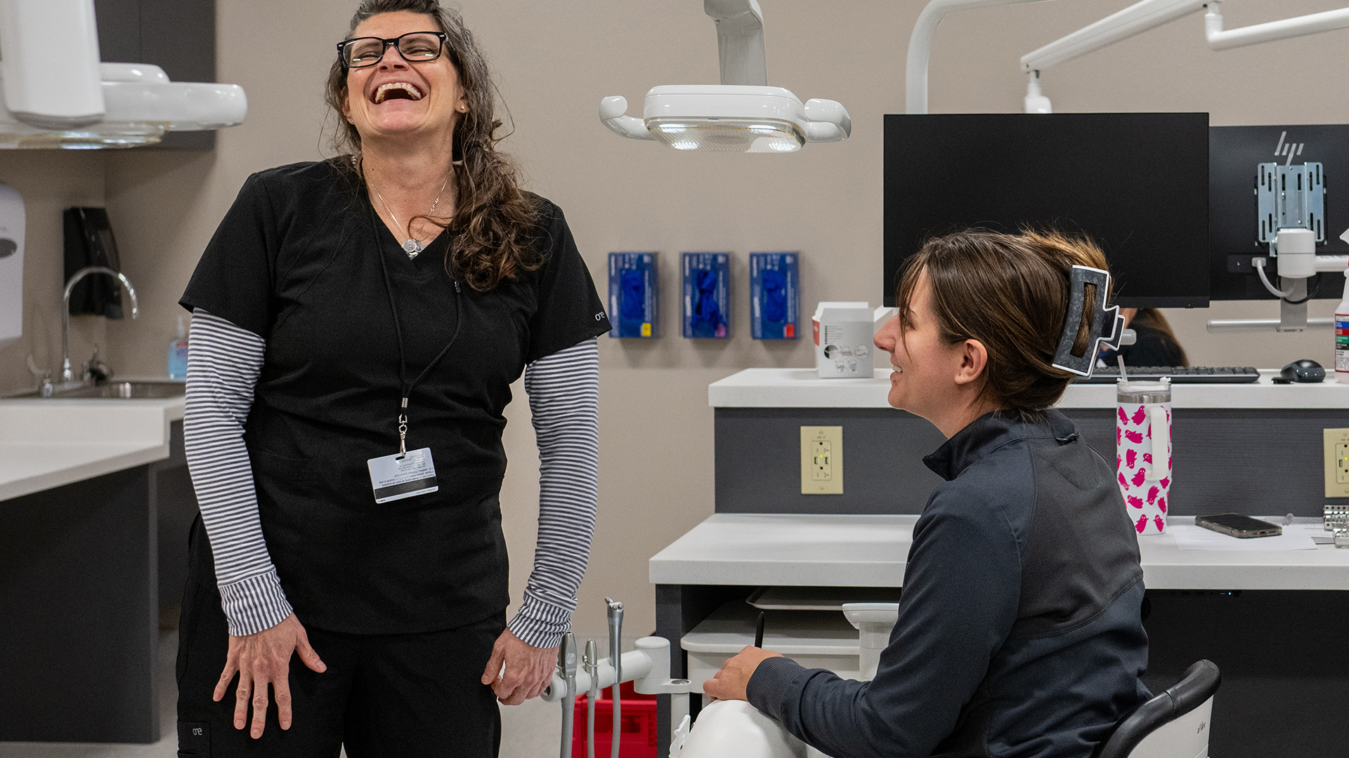 Robin Eichhorst laughs while standing next to Nikky K., who is seated in a rolling chair next to a multi-level table with multiple electrical sockets and a monitor on its upper surface, in a room with multiple adjustable dental lamps on arms overhead and a sink in a corner.
