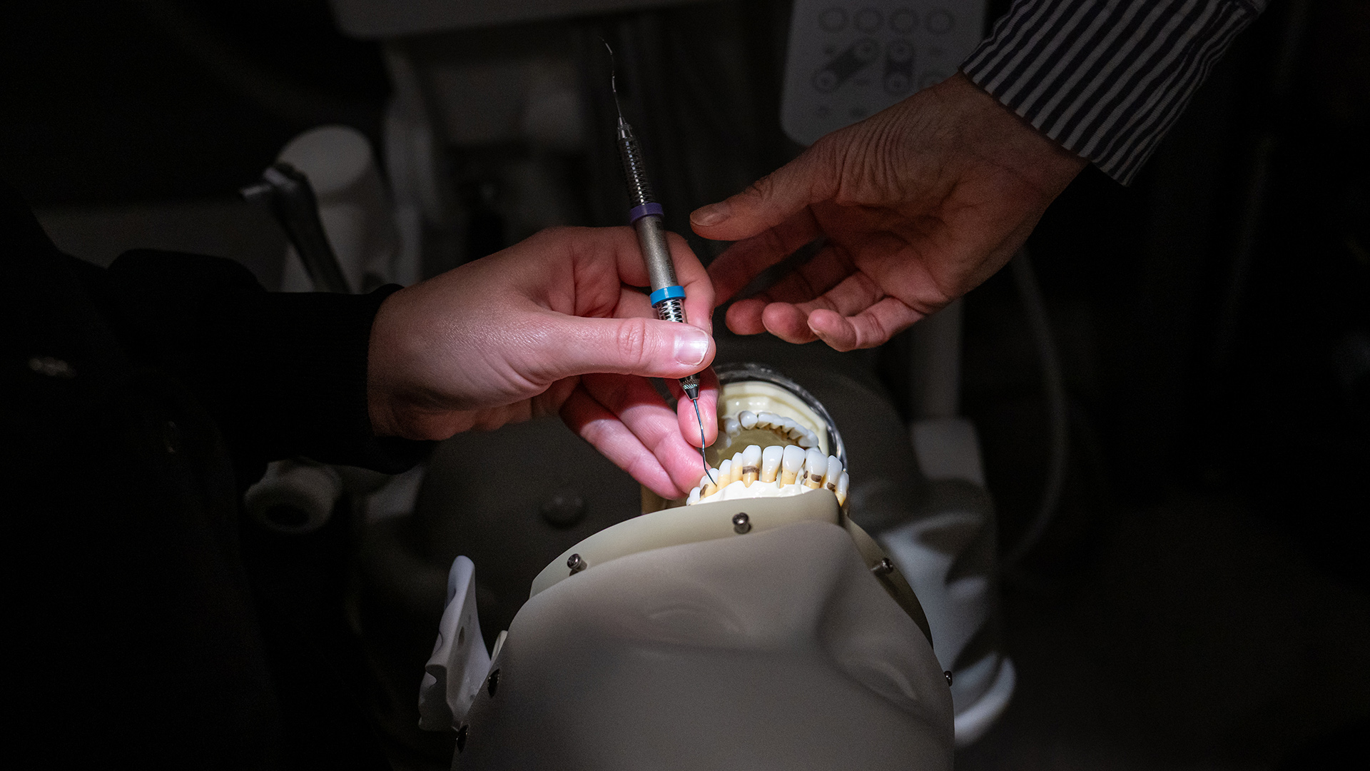 The right hand of Robin Eichhorst reaches toward the left hand of a student holding a metal dental pick in the open mouth of a mannequin placed in a prone position, with light shining on its face.