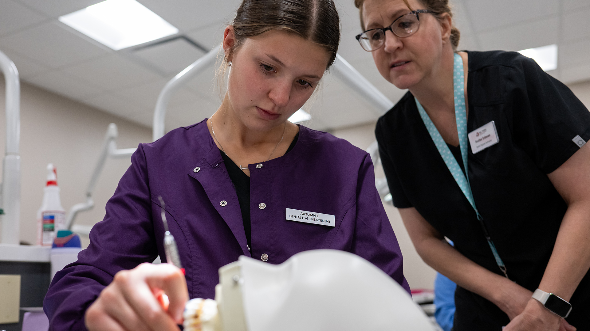 Autumn L. holds a metal dental pic in her right hand while practicing scaling teeth of an out-of-focus mannequin in the foreground, with Heather Erdmann standing behind her and watching in a room with a drop-panel ceiling and fluorescent lighting.