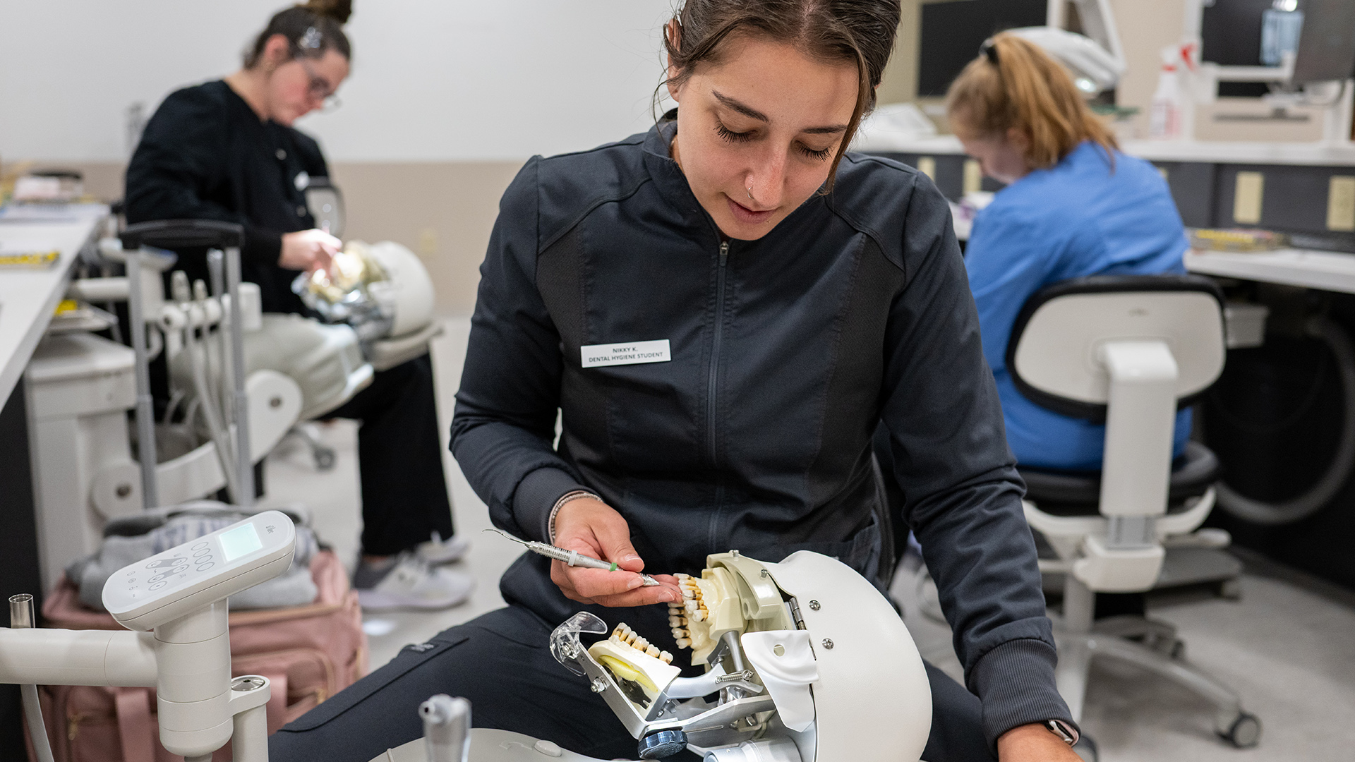 Nikky K. holds a metal dental pic in her right hand while practicing scaling teeth of a mannequin in the foreground, with two out-of-focus students seated in rolling chairs at other work stations in the background.