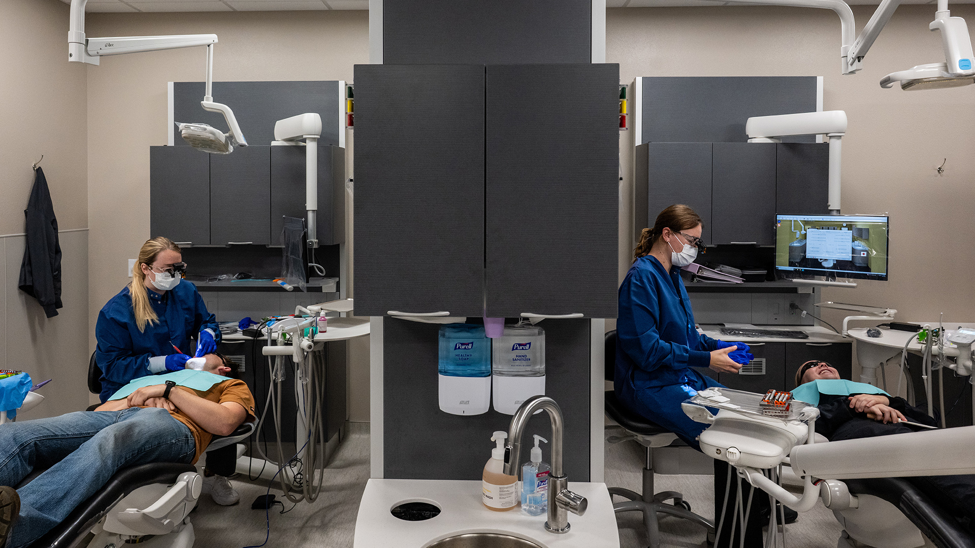 Kaitlyn Weyenberg and Kylie Konrad sit in rolling chairs while wearing scrubs, surgical masks and safety glasses with mounted lights next to patients in prone positions on dental chairs in two separate stations with adjustable dental lamps on arms overhead and shelves with cabinets in the background, with a sink with multiple dispensers of hand sanitizers and cabinets in the center in the foreground.