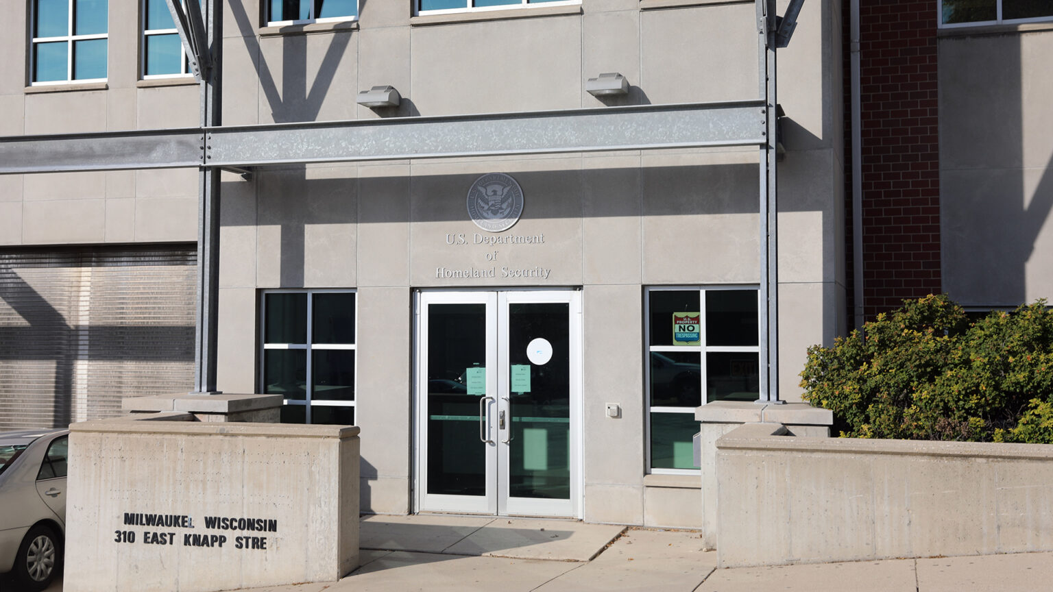 A car is parked next to a concrete wall with a letter sign on its front reading Milwauke Wisconsin and 310 East Knapp Stre that's situated along a sidewalk and in front of a multistory building with a stone and brick exterior and windows on either side of double glass doors below a letter sign reading U.S. Department of Homeland Security. A car is parked next to a concrete wall with a letter sign on its front reading Milwauke Wisconsin and 310 East Knapp Stre that's situated along a sidewalk and in front of a multistory building with a stone and brick exterior and windows on either side of double glass doors below a letter sign reading U.S. Department of Homeland Security.