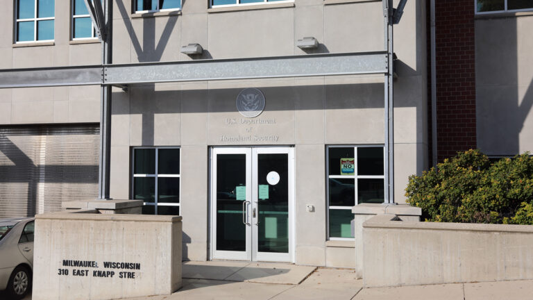 A car is parked next to a concrete wall with a letter sign on its front reading Milwauke Wisconsin and 310 East Knapp Stre that's situated along a sidewalk and in front of a multistory building with a stone and brick exterior and windows on either side of double glass doors below a letter sign reading U.S. Department of Homeland Security. A car is parked next to a concrete wall with a letter sign on its front reading Milwauke Wisconsin and 310 East Knapp Stre that's situated along a sidewalk and in front of a multistory building with a stone and brick exterior and windows on either side of double glass doors below a letter sign reading U.S. Department of Homeland Security.