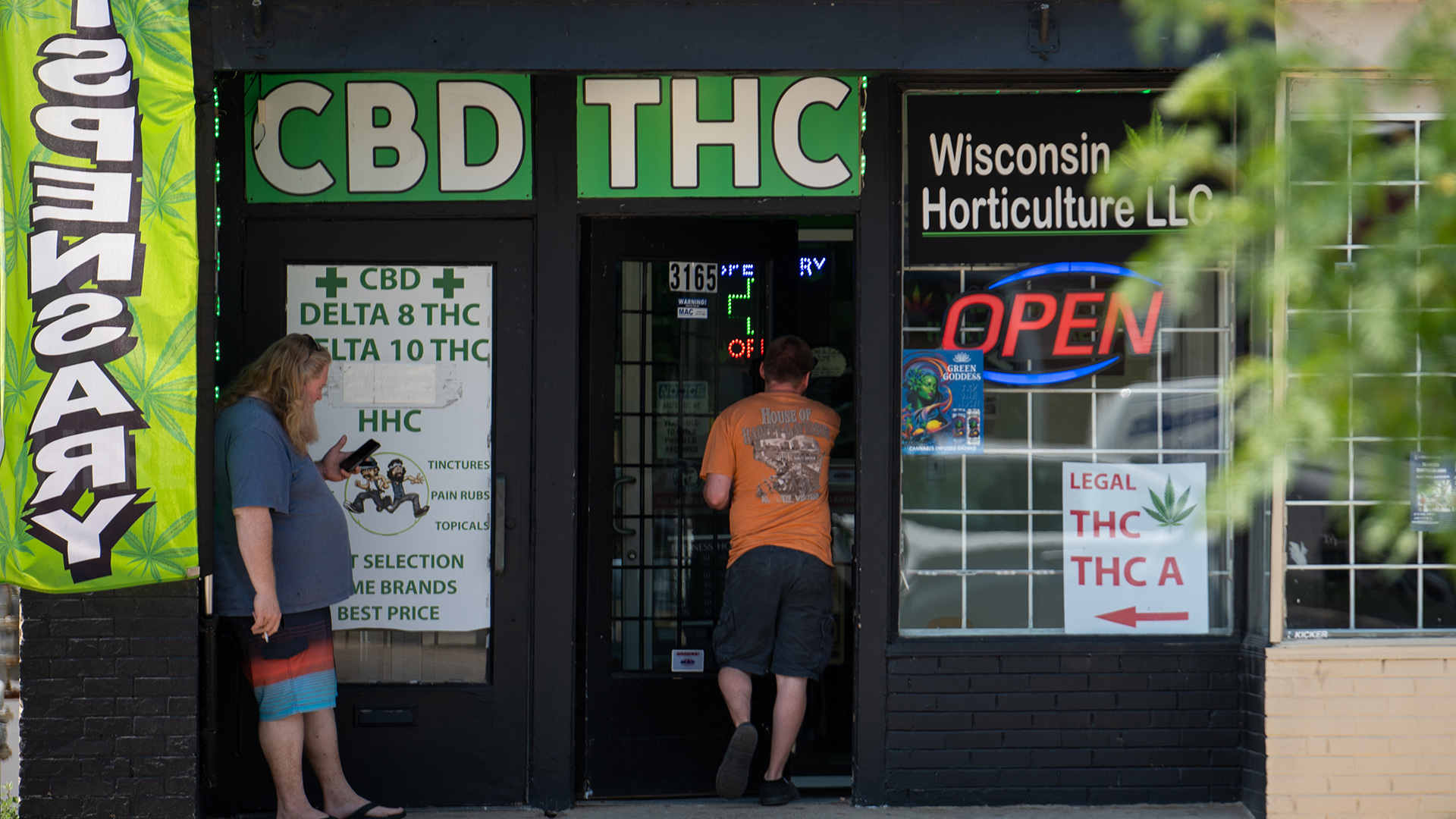 A person walks through the open door of a storefront of a painted brick building with multiple signs in windows reading "CBD," "THC," "CBD Delta 8 THC Delta 10 THC HHC," "Wisconsin Horticulture LLC," "Legal THC THC A" and a neon "Open" sign, with another person leaning against a wall next to the doorway and looking at a phone in their left hand.