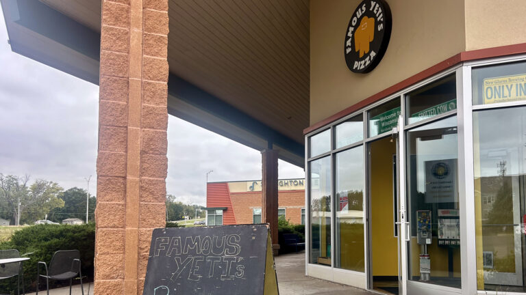 A sandwich board sign with the words Famous Yeti's written in chalk on its surface stands next to a brick pillar and in front of the entrance to a building with an open glass door and a wall of glass windows, below a round sign with a graphic of a cryptid and the words Famous Yeti's Pizza.