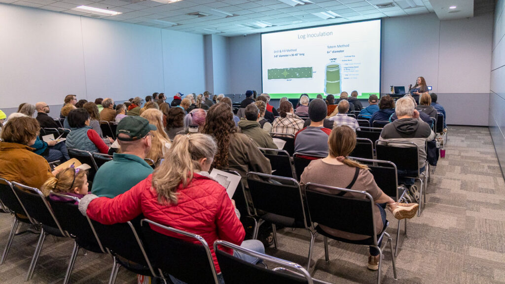 A presenter speaks to a crowded lecture room with a "Log Inoculation" slide projected on a screen.