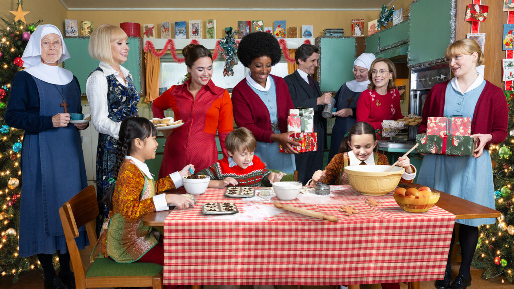 A group of midwives wearing nurse uniforms and sisters stand around a table where young children make Christmas desserts.
