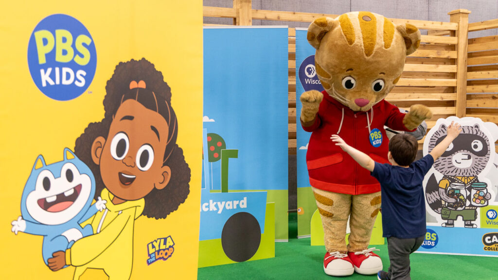 Daniel Tiger mascot greets a young child running his way in the PBS KIDS-themed area of an Expo center.