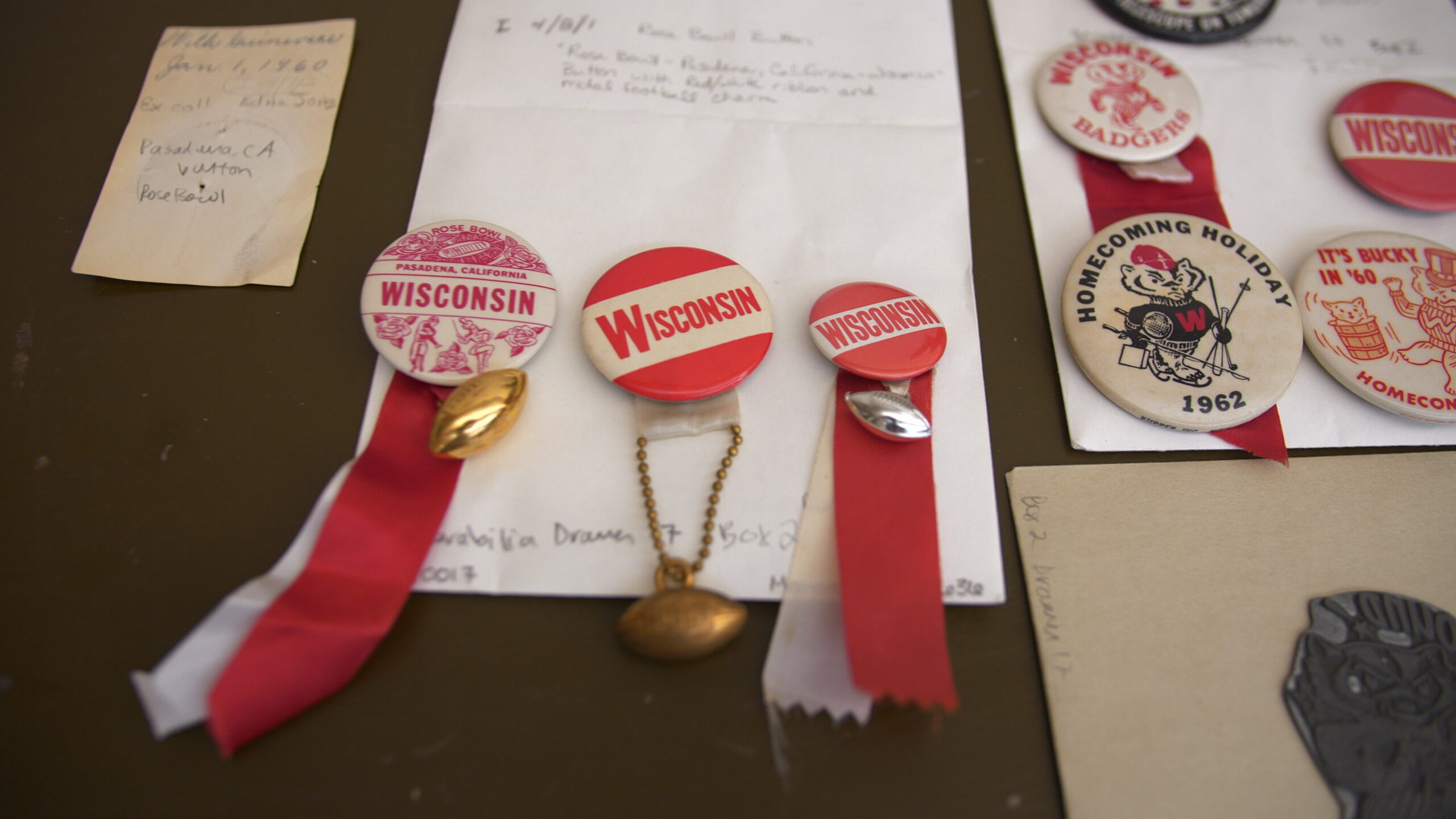 A collection of vintage Wisconsin Badgers memorabilia is displayed on a table. Items include buttons and badges with red ribbons, featuring "Wisconsin" text, mascots, and dates like 1962. The overall tone conveys nostalgia and school spirit.