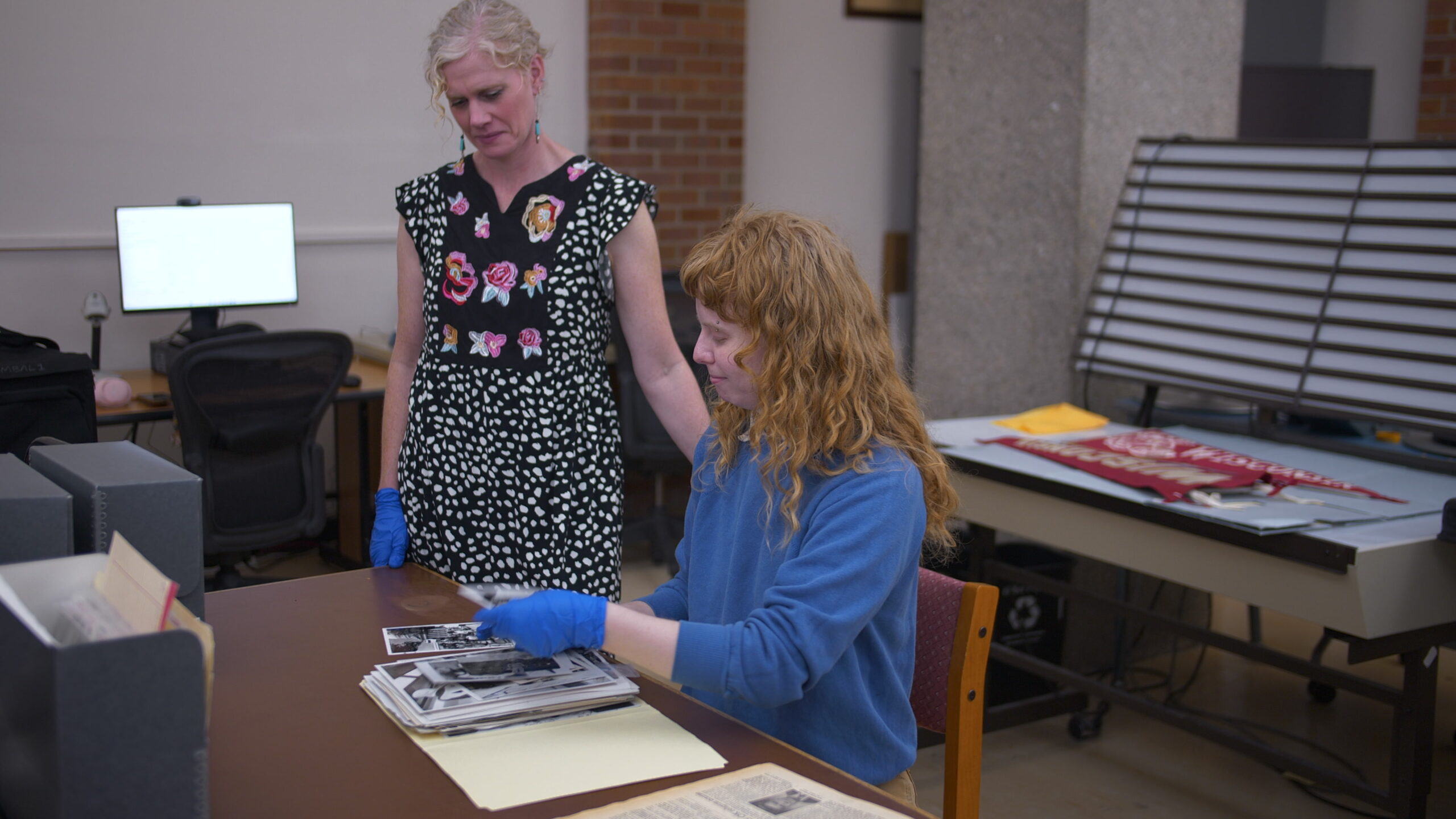UW-Madison Head of Archives Katie Nash (left) and PBS Wisconsin archivist Clara Wolfe (right) in a library or archive, one standing and one seated, both wearing blue gloves. They handle historical photos on a table, indicating careful preservation work.