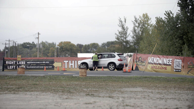 A person wearing a reflective safety vest stands in a driveway next to the open driver's window of a stopped vehicle among multiple traffic cones, in front of multiple vinyl advertising banners featuring the logo of a cannabis dispensary, the words Menominee and Welcome, a QR code, and directions for drivers, with utility poles and trees in the background.