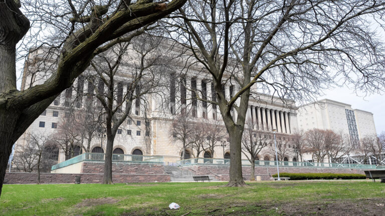 Multiple leafless trees stand on a lawn with lights, trash cans and park benches in front of a terraced plaza with brick walls and a weathered copper fence, with a multi-story masonry building with first-story arches and multiple colonnades at an upper level above two floors of windows. Multiple leafless trees stand on a lawn with lights, trash cans and park benches in front of a terraced plaza with brick walls and a weathered copper fence, with a multi-story masonry building with first-story arches and multiple colonnades at an upper level above two floors of windows.