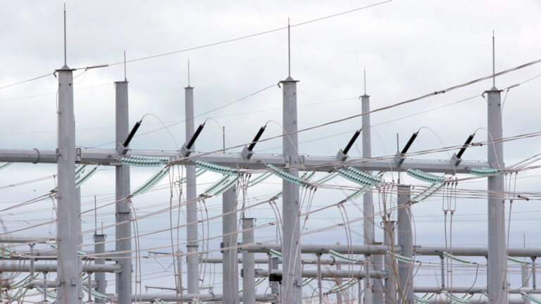 Multiple vertical towers and horizontal bars are connected by an array of multiple power lines, with portions of connecting lines covered by glass insulators, form part of an electrical substation, under a cloudy sky.