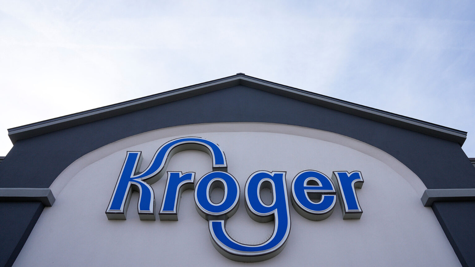 A letter sign of the word Kroger with stylized characters is attached to the face of a building with a peaked roof under a partly cloudy sky.