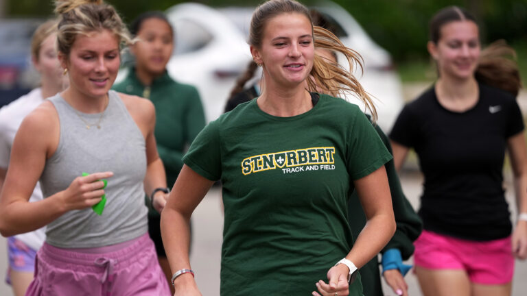 Madison DeCleene runs while wearing a t-shirt with a St. Norbert Track and Field graphic, with more than a half-dozen out-of-focus runners and a parked vehicle behind her. Madison DeCleene runs while wearing a t-shirt with a St. Norbert Track and Field graphic, with more than a half-dozen out-of-focus runners and a parked vehicle behind her.