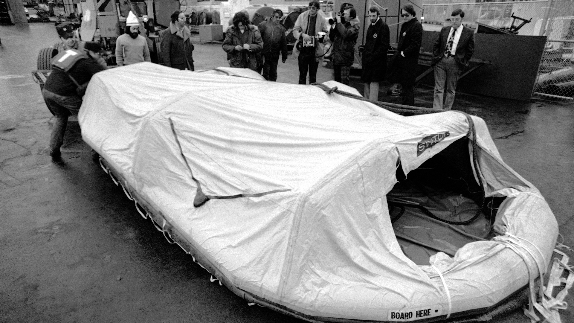 A grayscale photo shows an inflatable life raft with a cover and an opening at one end with a small sign reading "Board Here" being pulled along a concrete pad, with multiple people standing in the background in front of industrial equipment and a chain-link fence.