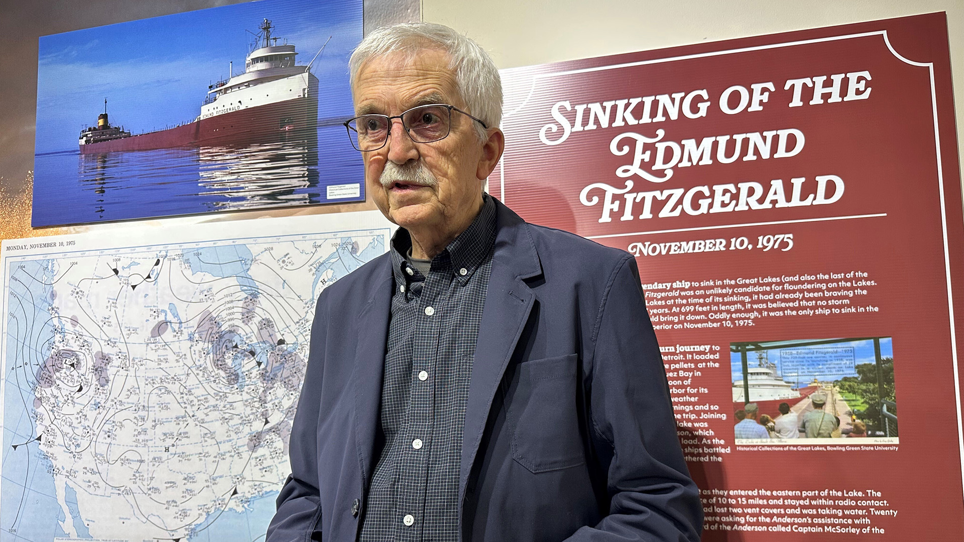 Frederick Stonehouse speaks while standing in front of a photo of the "Edmund Fitzgerald," a map of North America showing weather patterns, and an interpretive sign with the title text "Sinking of the Edmund Fitzgerald" and the date "November 10, 1975" followed by multiple paragraphs of text, each mounted on a wall and glazed glass window.